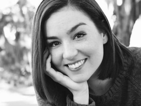 A casual black and white portrait of a smiling young woman with straight, shoulder length hair wearing a jumper, seated outdoors.