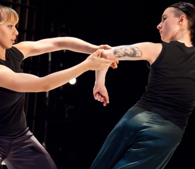 Two female dancers both dressed in black vests and trousers. One dancer is holding the arm of the other dancer who is leaning away and has a large tattoo of a female face on her armhas a
