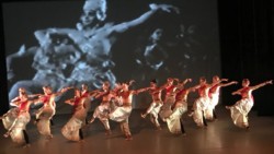 15 dancers in Indian costume performing onstage in front of black and white film projection