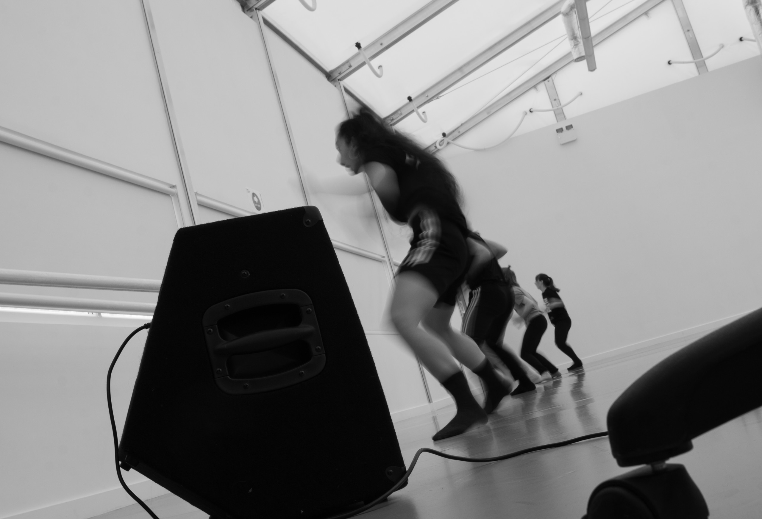 Dancers in a line at the back of the studio, there is a speaker in the foreground and they are rehearsing a dance piece