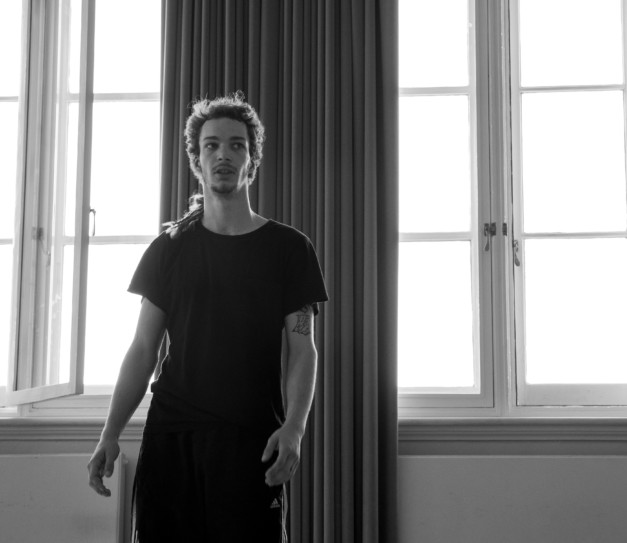A male dancer stands in a studio space, hands by his sides