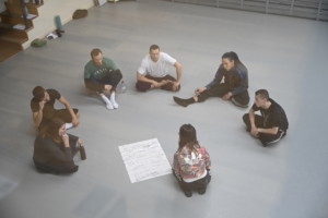 6 male dancers sat in circle in rehearsal room with choreographer Maxine