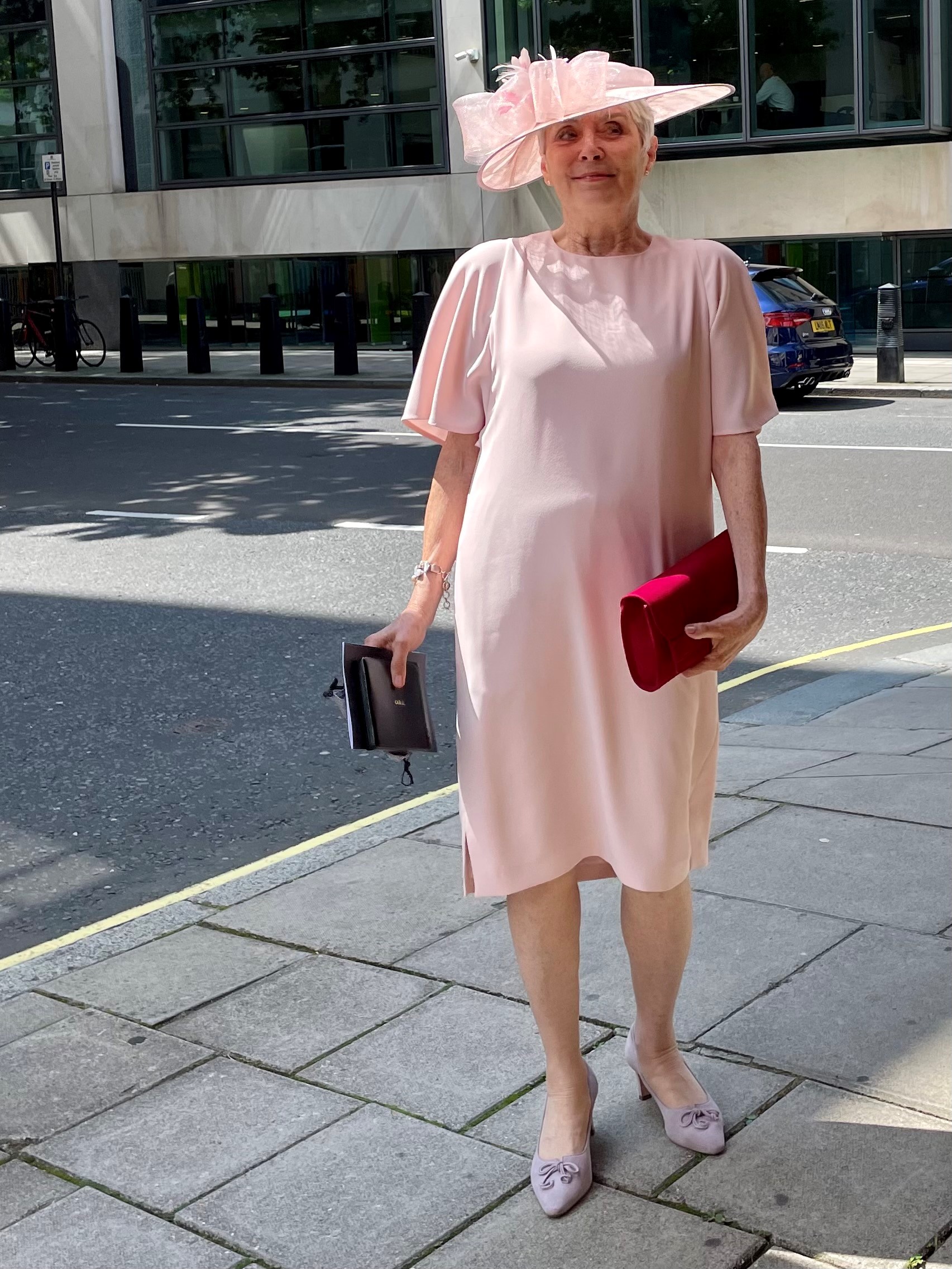 Mirella Bartrip OBE smiling in pink dress and hat with red clutch bag