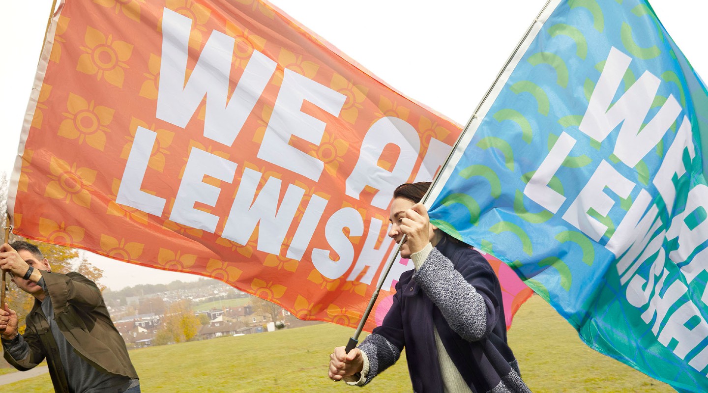 Man and woman running across green open space with large orange a blue 'We Are Lewisham' flags