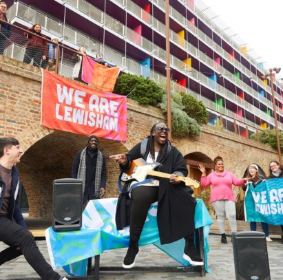 Dave Okumu playing guitar outside Deptford arches with dancers and branded 'We Are Lewisham' signs