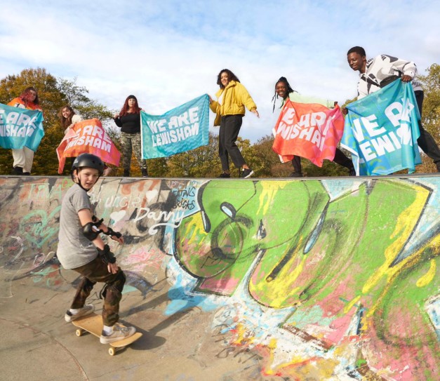 Skateboarders in graffitied outdoor skate park with 'We Are Lewisham' signs