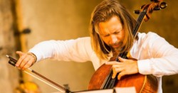 David Cohen with long hair and beard in white top, with arm raised holding bow, playing cello, looking down at fingerboard