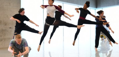Five young dancers in the studio leaping in the air with their legs at right angles and heads turned to the left, in the front ayoung male dancer is crouched down