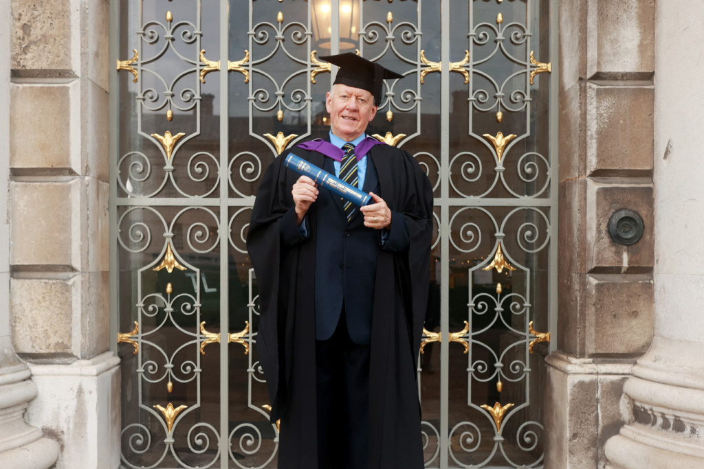 Philip Fowke standing in front of wrought iron gates and stonework in graduation hat and robe holding honorary fellowship scroll in both hands