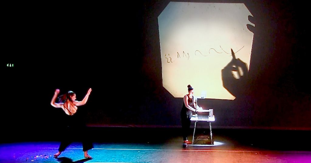 Performer documenting dancer's movement during live performance by using overhead projector showing markings on a page