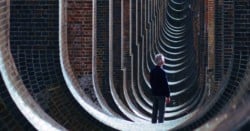 Nic Pendlebury standing in repeating arches of viaduct, facing away and looking into distance