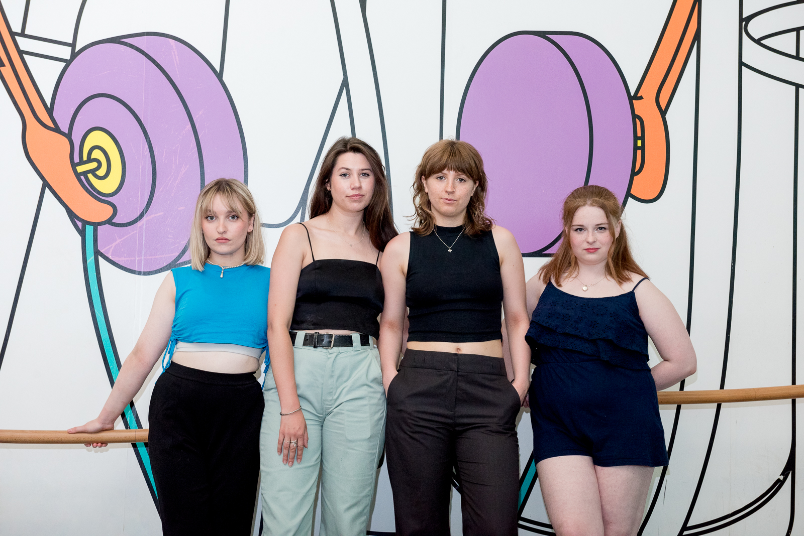 Phoebe Noble, Natasha Spencer Levy, Ellie Drayton and Holly McConville all leaning against a wooden bar with a coloured illustration on the wall behind them