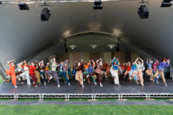 Mass Dance comes to Lewisham. Group of dancers in seven rows across a large open stage dancing with one leg raised to the knee and their hands above their head. They are wearing brightly coloured trousers and tops.