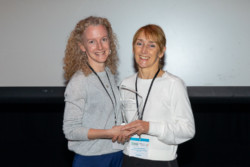 Edel Quinn presenting Sonia Rafferty with her IADMS Dance Educator award. Both are standing together against a white background, Both are smiling at the camera holding the award cupped in their hands.
