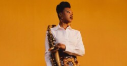 Camilla George stands against a bright orange background. She is wearing a white shirt and colourful skirt. She is holding her saxophone and looking to her left. Camilla has been nominated at the Parliamentary Jazz Awards 2023 for 'Jazz Instrumentalist of the Year'.