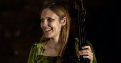 Valentina Ciardelli holds her double bass, she is wearing a green dress and stands in front of a black background.